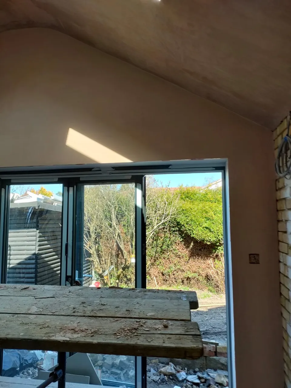 Plastered vaulted ceiling and walls beside black framed bifold doors, scaffold boards in the foreground